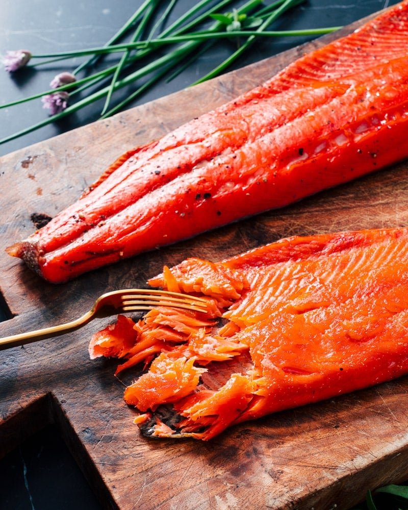 Two smoked salmon fillets on a cutting board, with one being shredded by a fork