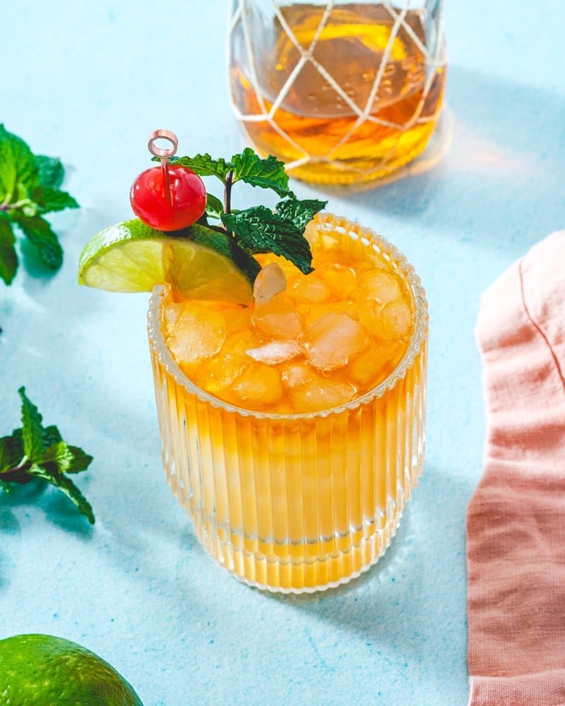 Mai Tai drink in a short glass with ice and garnished with lime, cherry, and fresh mint leaves