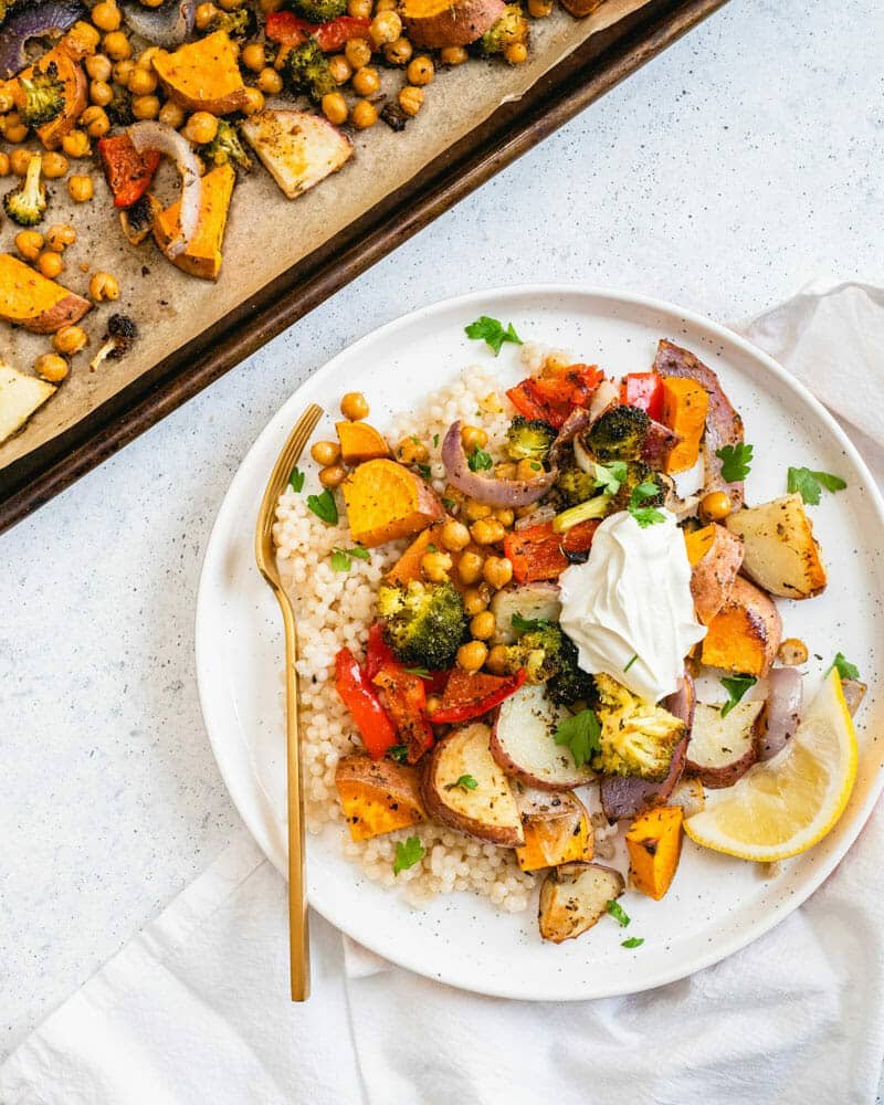 A sheet pan dinner on a plate served over couscous and topped with a mixture of veggies and chickpeas, and a lemon wedge for garnish