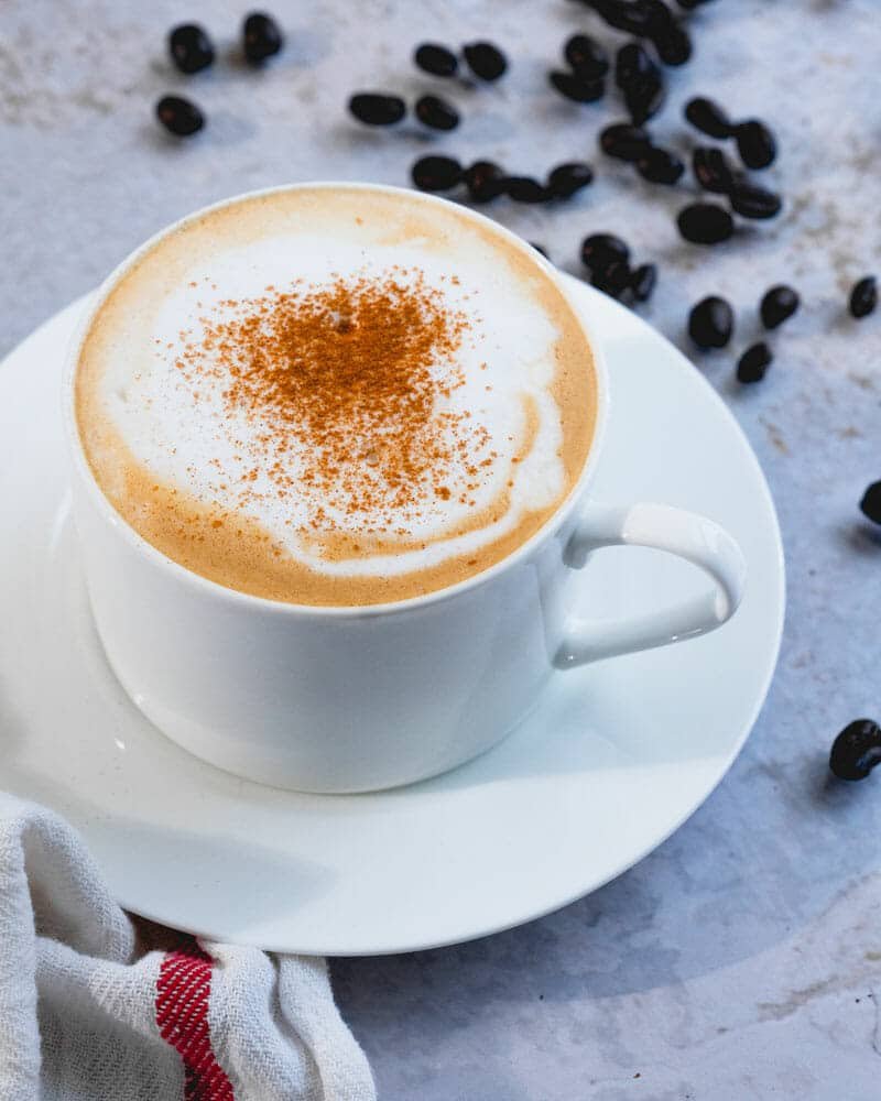 A cappuccino in a white cup and plate, surrounded by coffee beans, and sprinkled with cinnamon